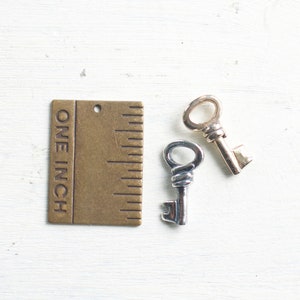 May include: Two small decorative keys, one silver and one gold, next to a brass ruler that reads "ONE INCH". The keys are likely for jewelry making or crafting. The keys are approximately 1 inch (2.54 cm) in length.