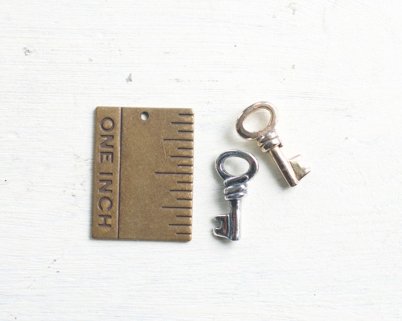 May include: Two small decorative keys, one silver and one gold, next to a brass ruler that reads "ONE INCH". The keys are likely for jewelry making or crafting. The keys are approximately 1 inch (2.54 cm) in length.