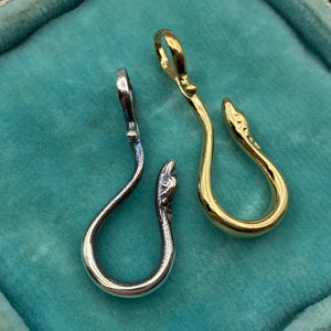 May include: Two snake-shaped pendants, one silver and one gold, on a turquoise velvet background. Both pendants are in the shape of a snake with its head forming a hook.