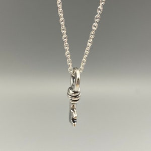 May include: A silver-colored pendant necklace featuring a stylized key design. The key pendant hangs from a delicate chain. The key has a detailed design, and the necklace is displayed against a neutral background.