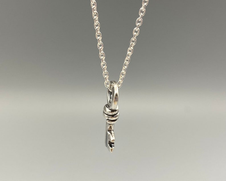 May include: A silver-colored pendant necklace featuring a stylized key design. The key pendant hangs from a delicate chain. The key has a detailed design, and the necklace is displayed against a neutral background.