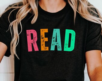 Puede incluir: Camiseta gris oscuro con la palabra "READ" en letras grandes, coloridas y desgastadas. Las letras son rosa, naranja, turquesa y verde. La camiseta es de estilo casual, adecuada para el uso diario.