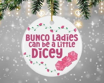 Puede incluir: Adorno redondo de cerámica blanca con el texto "Bunco Ladies can be a Little Dicey" en rosa. El adorno presenta dados rosas y detalles florales, ideal para un regalo de noche de juegos o decoración festiva.