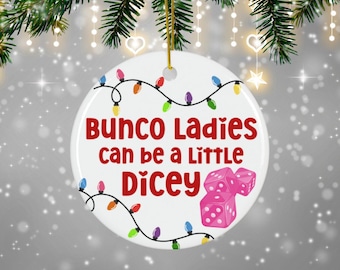 May include: A white ceramic ornament with the text "Bunco Ladies can be a Little Dicey" in red. The ornament features a string of colorful lights and pink dice. A festive holiday decoration.