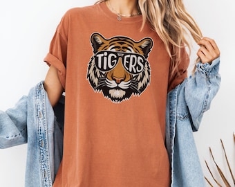 Puede incluir: Una camiseta de color óxido con un gráfico de un tigre con gafas de sol y la palabra "TIGERS" impresa en las gafas.