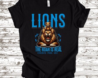 May include: Black t-shirt featuring a graphic of a roaring lion with a cub on its head. The word "LIONS" is above the lion graphic in blue, and the text "THE ROAR IS REAL" is below it. The shirt also has the words "STRENGTH. PRIDE. GRIT."