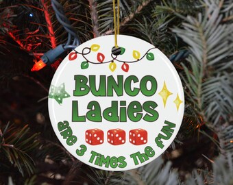 May include: A white, round Christmas ornament with the text "BUNCO Ladies are 3 Times The FUN" in green. The ornament features red dice, a green star, and yellow sparkles, with a string of colorful lights at the top.