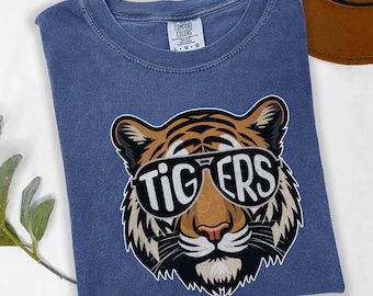 Puede incluir: Camiseta azul denim con un gráfico de un tigre con gafas de sol que dice "TIGERS". La camiseta es de la marca Comfort Colors, con cuello redondo y mangas cortas. La camiseta está doblada.