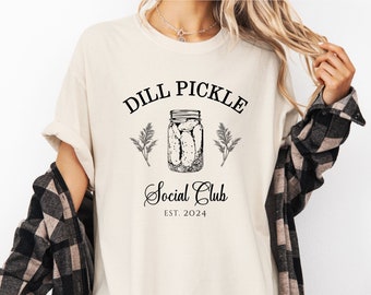 May include: Cream-colored t-shirt with a black graphic that reads "DILL PICKLE Social Club EST. 2024." The design includes a jar of pickles and sprigs of greenery. The shirt has a relaxed fit.