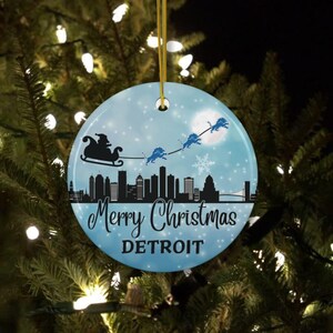 May include: A blue and white round ceramic Christmas ornament featuring a black silhouette of the Detroit skyline with Santa Claus and reindeer flying over the city. The ornament reads "Merry Christmas DETROIT".