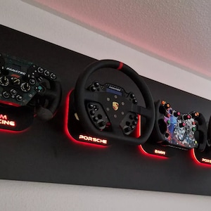 Könnte beinhalten: Eine schwarze Wandhalterung mit vier Lenkrädern. Die Lenkräder sind auf einem schwarzen Hintergrund mit roter LED-Beleuchtung montiert. Die Lenkräder sind mit "Sim Racing", "Porsche", "BMW" und "Porsche" beschriftet.