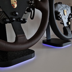 Könnte beinhalten: Vier schwarze Lenkräder mit unterschiedlichen Designs werden auf schwarzen Ständern mit blauen LED-Lichtern präsentiert. Die Lenkräder haben braunes Leder, schwarze und weiße Akzente und ein Porsche-Logo.