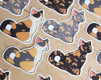 May include: A collection of cat stickers in various calico patterns. The stickers feature cats in shades of black, orange, gray, and white, with pink-lined ears. The stickers are arranged on a light brown wooden surface.