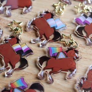 May include: Acrylic keychains featuring a brown horse with a black mane and tail, holding a flag with various LGBTQ+ pride flag designs. Each keychain has a gold star-shaped clasp.