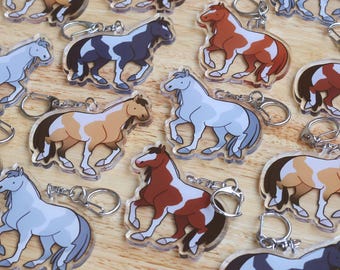 Puede incluir: Una colección de llaveros con forma de caballo en varios colores, incluyendo gris, marrón y negro, con llaveros plateados. Los llaveros están dispuestos sobre una superficie de madera marrón claro.
