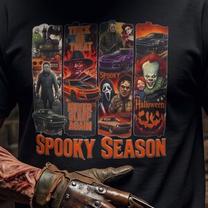 May include: A black t-shirt featuring a graphic print with multiple horror movie characters, cars, and the text "TRICK OR TREAT," "NEVER SLEEP AGAIN," "SPOOKY," "HALLOWEEN," and "SPOOKY SEASON" in orange and red tones on the front chest area.