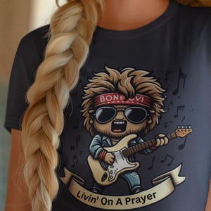 Könnte beinhalten: Ein dunkelblaues T-Shirt mit einer Cartoon-Illustration eines Rockstars, der Gitarre spielt. Die Illustration ist von Noten und einem Banner mit der Aufschrift "Livin' On A Prayer" umgeben.