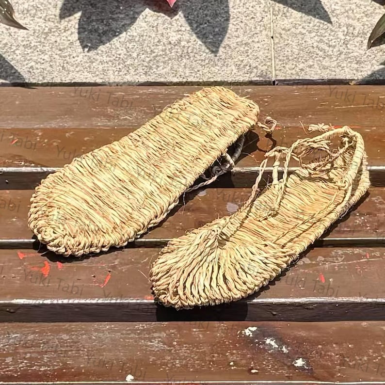 Op de afbeelding: Een paar traditionele Japanse strooien sandalen, of waraji, gemaakt van geweven stro. De sandalen zijn lichtbruin van kleur en hebben een eenvoudig ontwerp met bandjes om aan de voet te bevestigen. De sandalen worden op een houten oppervlak weergegeven.