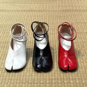 May include: Three pairs of Tabi shoes in white, black, and red. Each shoe has a split toe design and ankle straps. The shoes are displayed on a woven mat, showcasing their unique style and design.