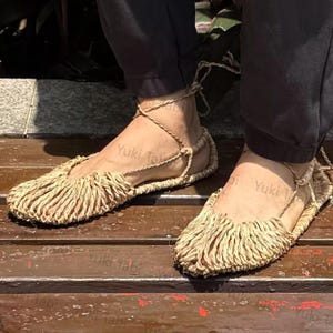 Op de afbeelding: Een paar sandalen van natuurlijke vezels met een gefranjerd teenontwerp. De geweven sandalen worden vastgezet met enkelbanden. Het schoeisel wordt gepresenteerd op een houten oppervlak, wat een rustieke esthetiek laat zien.
