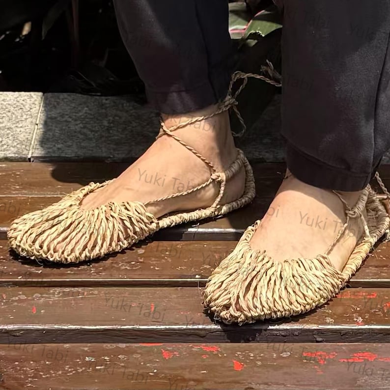 Op de afbeelding: Een paar sandalen van natuurlijke vezels met een gefranjerd teenontwerp. De geweven sandalen worden vastgezet met enkelbanden. Het schoeisel wordt gepresenteerd op een houten oppervlak, wat een rustieke esthetiek laat zien.