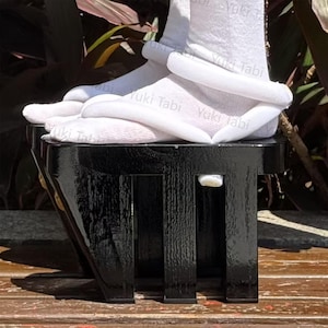 Puede incluir: Sandalias geta de madera lacada en negro con base rectangular y tres soportes verticales. Calcetines tabi blancos con el texto "Yuki Tabi" se usan con las geta. Las sandalias están sobre una superficie de madera.