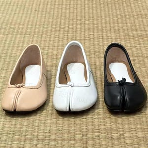 Op de afbeelding: Drie paar split-teen ballerina's in lichtbeige, wit en zwart. Elke schoen heeft een ronde neus en een klein knoopdetail. De schoenen staan op een geweven mat.