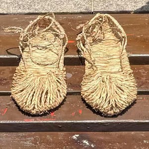 Op de afbeelding: Een paar traditionele Japanse strooien sandalen, of waraji, met een geweven ontwerp. De sandalen zijn lichtbruin en hebben een gefranjerde teen en geweven bandjes. Ze liggen op een houten oppervlak.