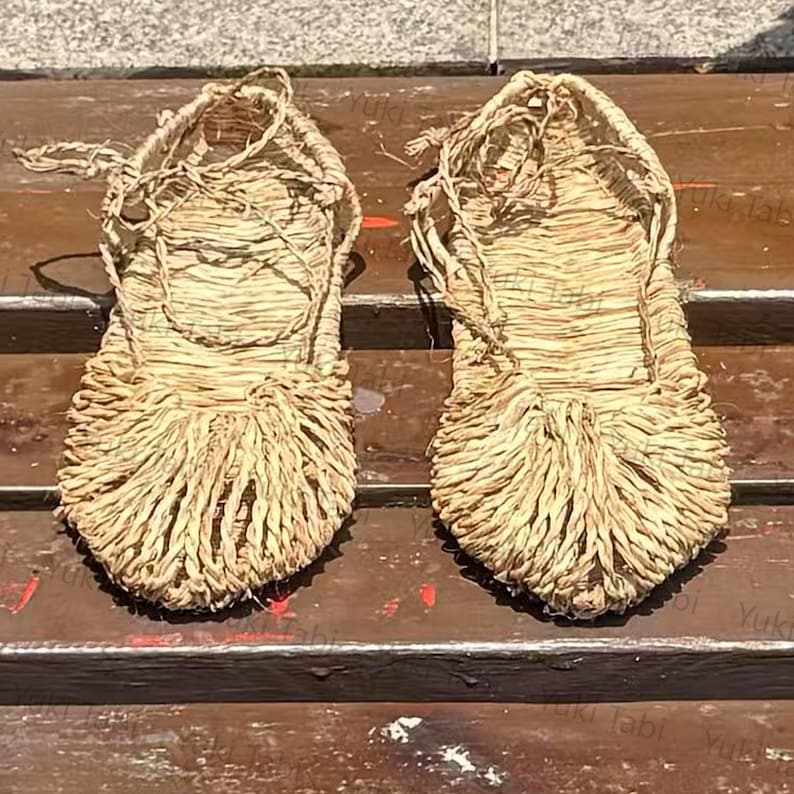 Op de afbeelding: Een paar traditionele Japanse strooien sandalen, of waraji, met een geweven ontwerp. De sandalen zijn lichtbruin en hebben een gefranjerde teen en geweven bandjes. Ze liggen op een houten oppervlak.