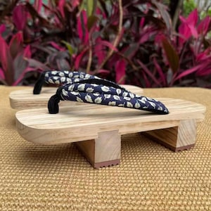 May include: A pair of traditional Japanese geta sandals. The wooden platforms are light-colored, with a curved front and two wooden supports. The straps are navy blue with a white leaf pattern and black thongs.