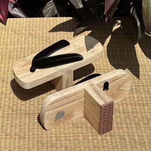 Pode incluir: Um par de sandálias geta japonesas tradicionais. As sandálias de madeira têm um acabamento de madeira natural com tiras de tecido preto. As solas têm uma aderência de borracha castanha. As sandálias são exibidas em uma esteira tecida.