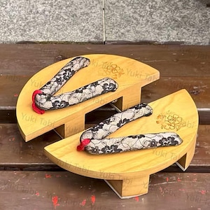 Puede incluir: Un par de sandalias geta de madera con correas de encaje floral negro y detalles rojos. Las sandalias tienen un acabado de madera natural y están elevadas sobre pequeños bloques de madera. El texto "Yuki Tabi" está presente.