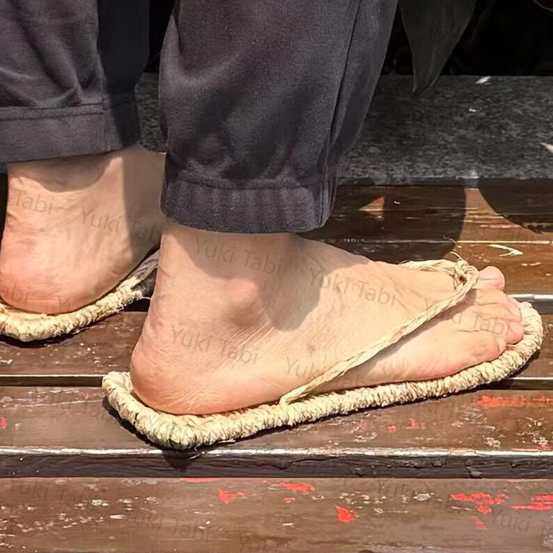 May include: Close-up of traditional Japanese straw sandals, or waraji, with a woven sole and a thong-like strap. The sandals are worn on a wooden surface. The text "Yuki Tabi" is visible on the sandals.