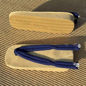 Può includere: Due paia di sandali Yuki Tabi. Le suole sono beige con un motivo a zig-zag. I cinturini sono blu navy e imbottiti. I plantari sono beige con la scritta "Yuki Tabi". Su una superficie tessuta marrone.
