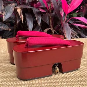 May include: A pair of traditional Japanese geta sandals. The wooden platform sandals are a deep red color with a glossy finish. The straps are a vibrant magenta, and a small gold bell hangs from the front of one sandal. The background features lush green and pink foliage.