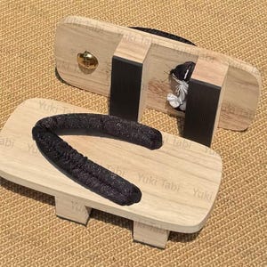 May include: A pair of traditional Japanese geta sandals. The wooden platforms are light-colored with black straps and a gold-colored bell. The sandals are designed for comfort and are a part of Japanese culture.