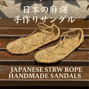 Op de afbeelding: Paar handgemaakte Japanse sandalen van stro touw. De sandalen zijn lichtbruin en hebben een geweven ontwerp. De afbeelding bevat Japanse tekst en de Engelse tekst "JAPANESE STRW ROPE HANDMADE SANDALS".