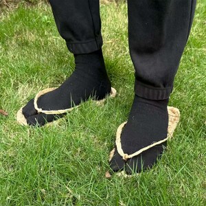 May include: Black socks and traditional Japanese straw sandals, known as geta, are worn on a grassy surface. The geta have a natural straw color and a thong that separates the big toe from the other toes. The person is wearing black pants.