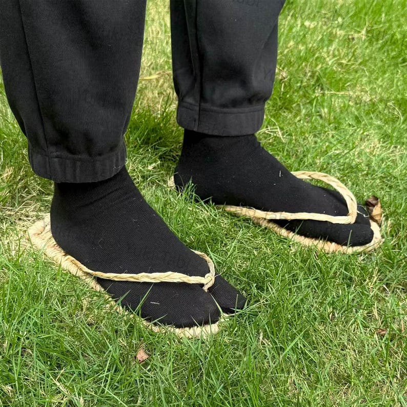 May include: A person wearing black socks and traditional Japanese straw sandals, known as geta, on a grassy surface. The geta feature a woven straw base and a thong that separates the big toe from the other toes.