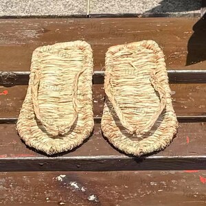 May include: A pair of traditional Japanese straw sandals, or waraji, with a woven design. The sandals are light brown and feature a thong-style strap. The image is taken from a high angle, showcasing the texture and construction of the footwear.