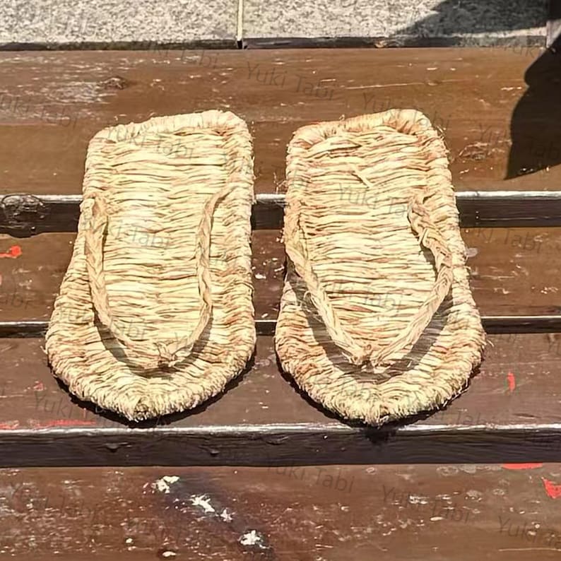 May include: A pair of traditional Japanese straw sandals, or waraji, with a woven design. The sandals are light brown and feature a thong-style strap. The image is taken from a high angle, showcasing the texture and construction of the footwear.