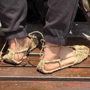 Op de afbeelding: Close-up van traditionele Japanse strooien sandalen, of waraji, gedragen op voeten. De sandalen zijn gemaakt van geweven stro en vastgezet met touw. De persoon draagt een zwarte broek.