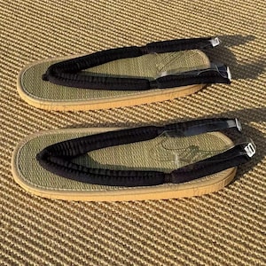 Può includere: Un paio di sandali geta giapponesi tradizionali. I sandali presentano un plantare intrecciato verde chiaro, una suola beige e cinturini in tessuto nero. I cinturini sono fissati con piccole fibbie metalliche.