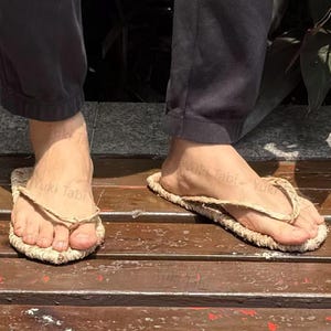 May include: A pair of natural-colored straw sandals with a thong-style design. The sandals are worn on a wooden surface, showcasing their simple, earthy aesthetic. The text "Yuki Tabi" is visible on the sandals.