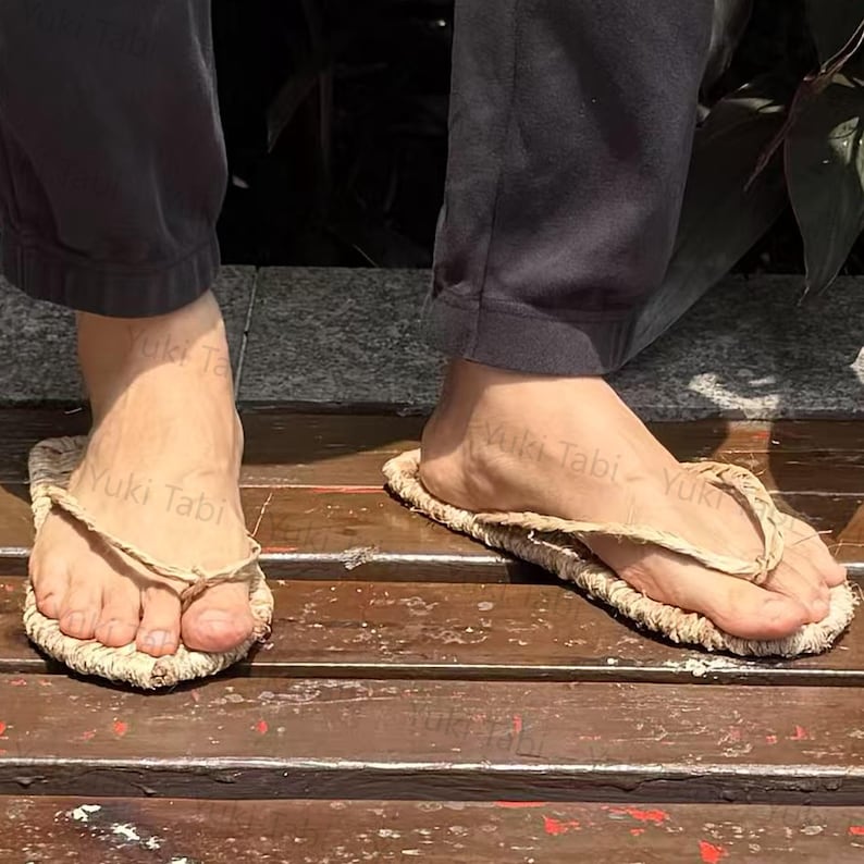 May include: A pair of natural-colored straw sandals with a thong-style design. The sandals are worn on a wooden surface, showcasing their simple, earthy aesthetic. The text "Yuki Tabi" is visible on the sandals.