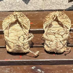 Op de afbeelding: Een paar traditionele Japanse strooien sandalen, of waraji, wordt getoond. De sandalen zijn gemaakt van geweven stro, met een afgeronde teen en een band om de voet vast te zetten. De natuurlijke beige kleur van het stro is prominent aanwezig, met zichtbare texturen en patronen van het weven.