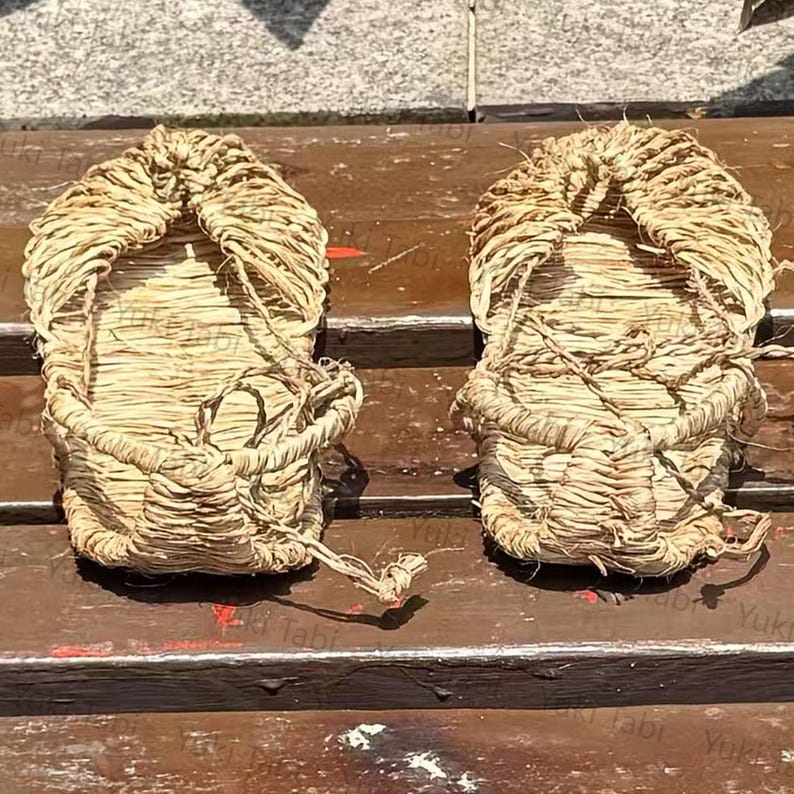 Op de afbeelding: Een paar traditionele Japanse strooien sandalen, of waraji, wordt getoond. De sandalen zijn gemaakt van geweven stro, met een afgeronde teen en een band om de voet vast te zetten. De natuurlijke beige kleur van het stro is prominent aanwezig, met zichtbare texturen en patronen van het weven.