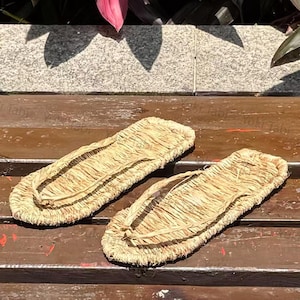 May include: A pair of traditional Japanese straw sandals, or waraji, are displayed. The sandals are light brown, rectangular, and feature a thong-style strap. They are made of woven straw and rest on a wooden surface.