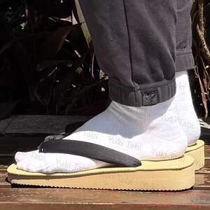 Può includere: Una persona indossa calzini bianchi e sandali geta giapponesi tradizionali. I sandali hanno una base marrone chiaro con un cinturino nero. Il testo "Yuki Tabi" è visibile sui calzini.