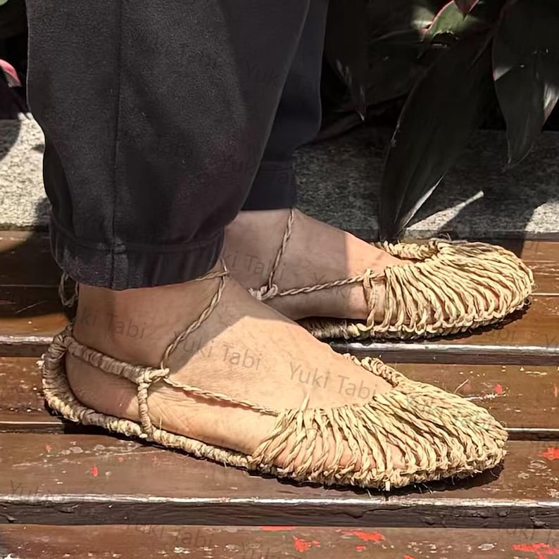 Op de afbeelding: Handgemaakte sandalen van natuurlijke vezels met een gefranjerde teen. De sandalen hebben enkelbandjes en zijn gemaakt van een lichtbruin geweven materiaal. De afbeelding toont de sandalen aan de voeten van een persoon.
